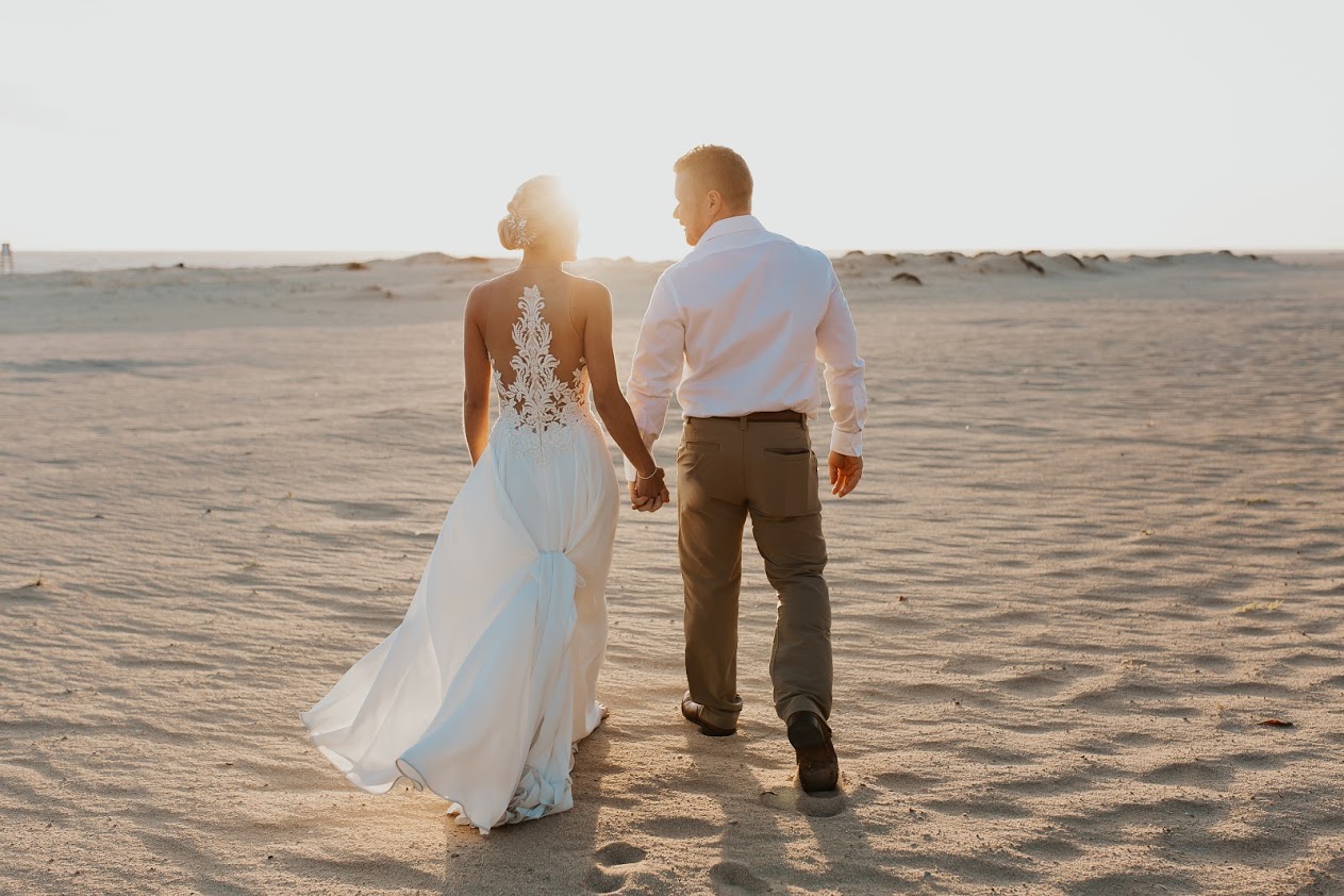 Bride in white dress groom in white shirt and tan pants walk on beach 
