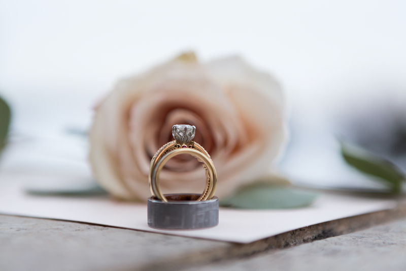 Gold engagement ring and black wedding ring sit in front of pink rose