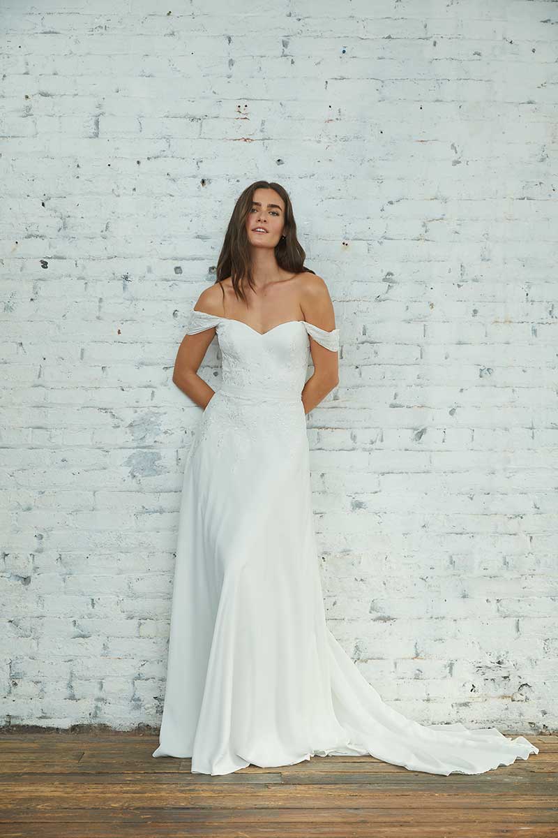 Bride in off the shoulder gown stands against white brick wall