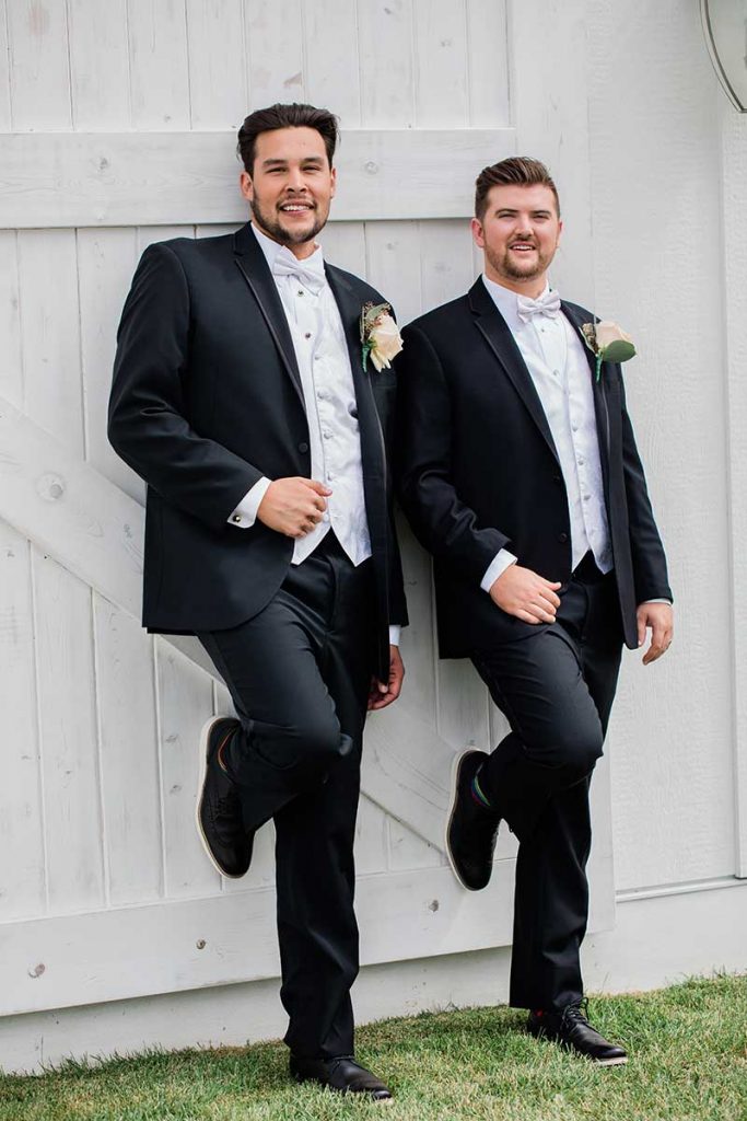 Two grooms in black suits lean up against a white barn door