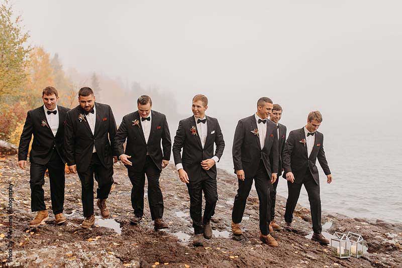 Groomsmen and groom in black suits walk along rocks on the North Shore in Minnesota