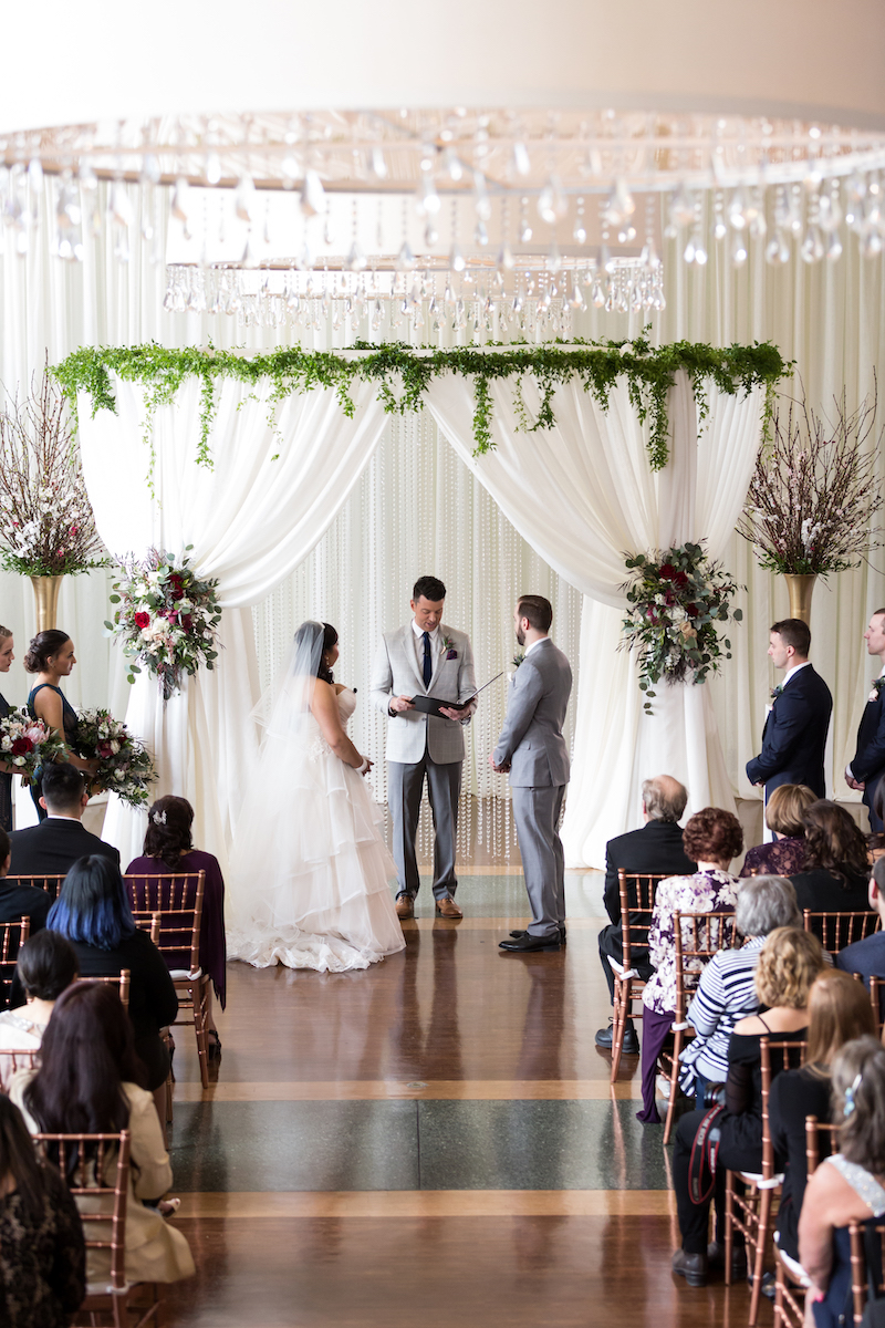 Couple stands under white draped arch with greenery