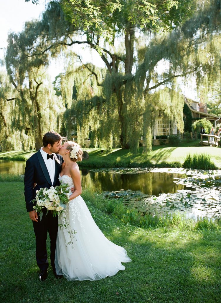 Bride and groom kissing outside near pond. Bride wears strapless Mira Zwillinger gown outdoor wedding willow tree