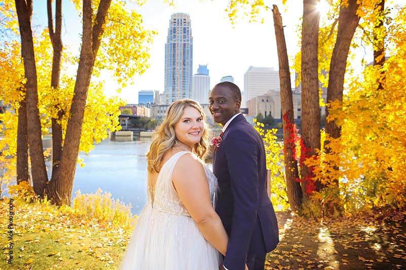 Bride and groom stand between fall trees and Minneapolis skyline