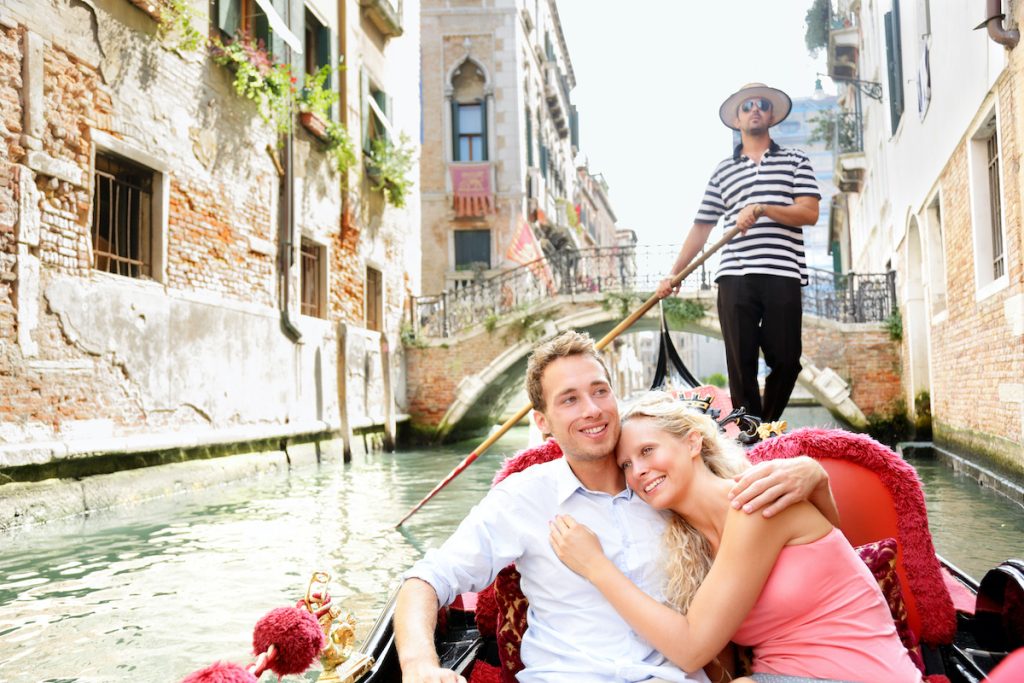 Romantic travel couple in Venice on Gondole ride romance in boat happy together on travel vacation holidays. Romantic young beautiful couple sailing in venetian canal in gondola. Italy, Europe planned by Travel Leaders