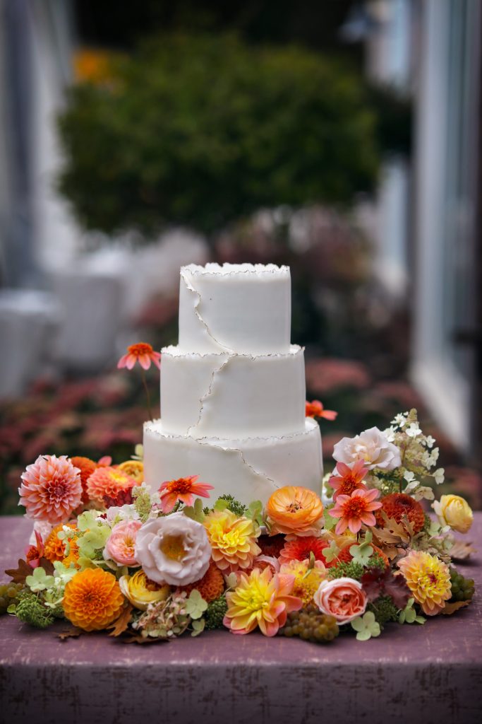 3-tier white wedding cake with red, orange, and yellow flowers surrounding it